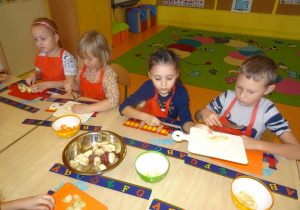 Czworo dzieci siedzi przy stole nakrytym podkładkami z rozłożonymi deskami, w ręku trzymają noże, którymi kroją owoce.